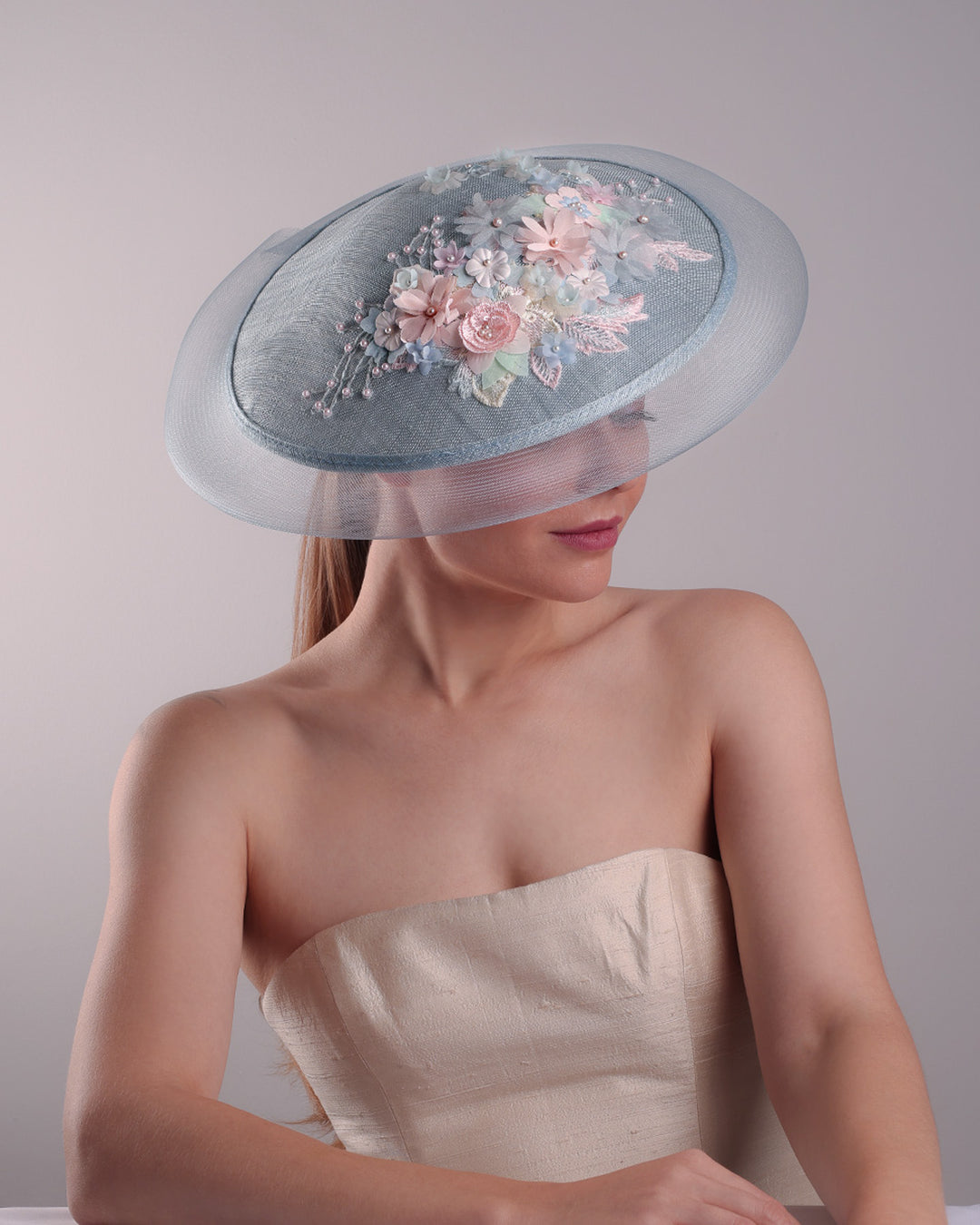 Model is showcasing the top view of a blue circle style saucer hat, extended with crinoline, decorated on the top with a 3D embroidery that is embellised with pearls, and silk flowers. The hat is worn tilted to the right side. The hat is fitted to the head with a wire headband and elastic.