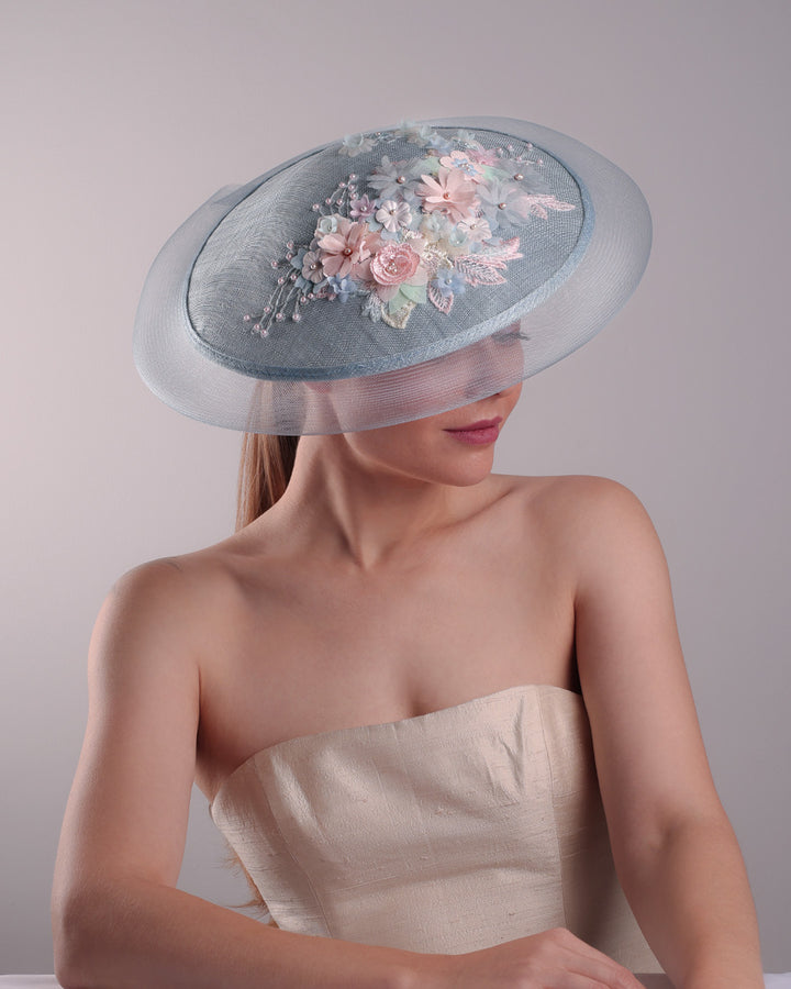 Model is showcasing the top view of a blue circle style saucer hat, extended with crinoline, decorated on the top with a 3D embroidery that is embellised with pearls, and silk flowers. The hat is worn tilted to the right side. The hat is fitted to the head with a wire headband and elastic.