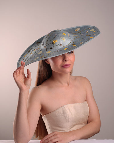 The model is showcasing the right angle of a blue asymetric whide brim hat, tilted to the right, decorated with pressed natural flowers creatinga almost 3D dimensional print affect on the hat. Fitted with a wired headband and additionally secured with elastic.
