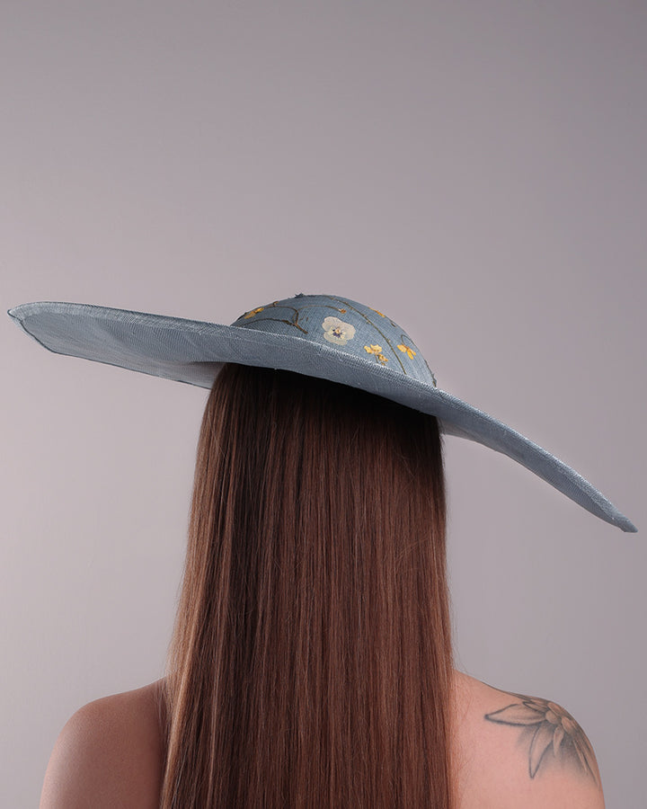 The model is showcasing the back side of a blue asymetric whide brim hat, tilted to the right, decorated with pressed natural flowers creatinga almost 3D dimensional print affect on the hat. Fitted with a wired headband and additionally secured with elastic.
