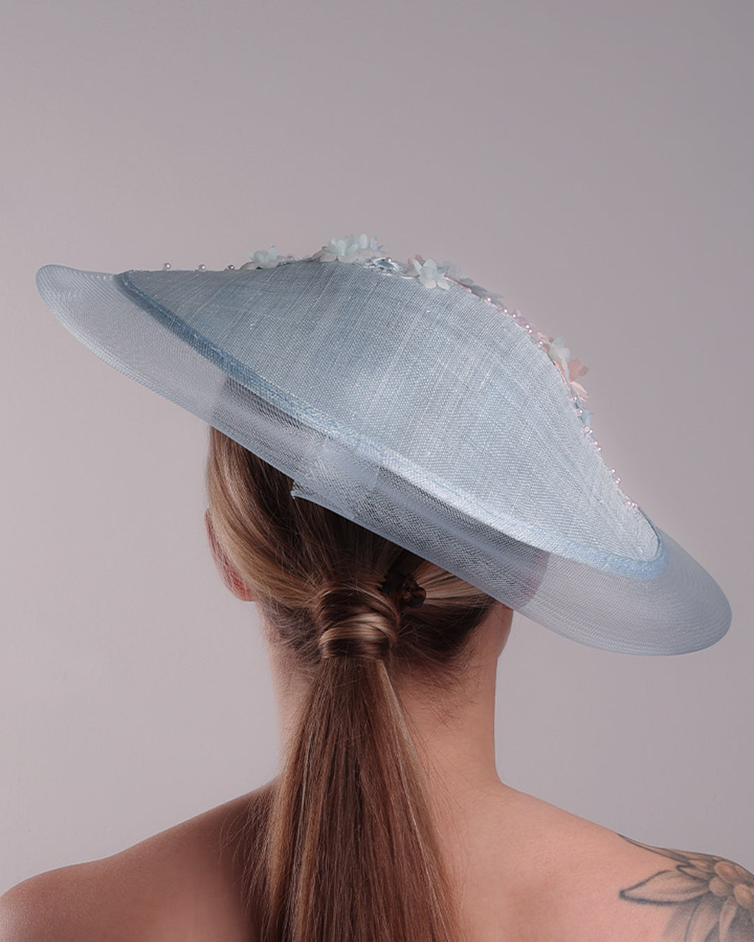 Model is showcasing the back of a blue circle style saucer hat, extended with crinoline, decorated on the top with a 3D embroidery that is embellised with pearls, and silk flowers. The hat is worn tilted to the right side. The hat is fitted to the head with a wire headband and elastic.