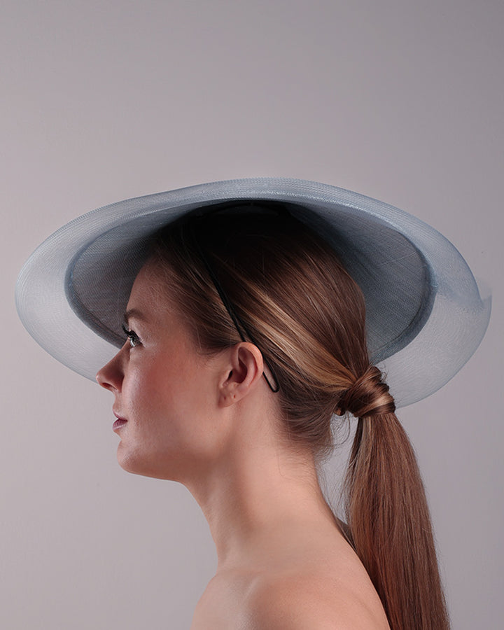 Model is showcasing the left side of a blue circle style saucer hat, extended with crinoline, decorated on the top with a 3D embroidery that is embellised with pearls, and silk flowers. The hat is worn tilted to the right side. The hat is fitted to the head with a wire headband and elastic.