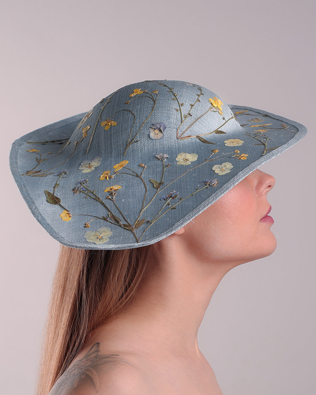 The model is showcasing the right side of a blue asymetric whide brim hat, tilted to the right, decorated with pressed natural flowers creatinga almost 3D dimensional print affect on the hat. Fitted with a wired headband and additionally secured with elastic.
