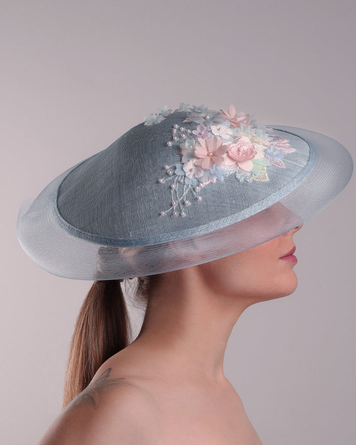 Model is showcasing the right side of a blue circle style saucer hat, extended with crinoline, decorated on the top with a 3D embroidery that is embellised with pearls, and silk flowers. The hat is worn tilted to the right side. The hat is fitted to the head with a wire headband and elastic.