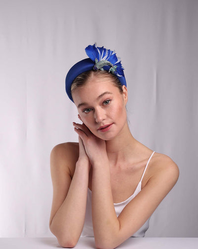 The model wears a cobalt blue high headband decorated with blue shades of crystal-adorned koi fish and shades of feathers behind the fish on the left side of the headband, creating an illusion of a wave. The high-fashion contemporary headband is something easy to wear yet makes a statement. 