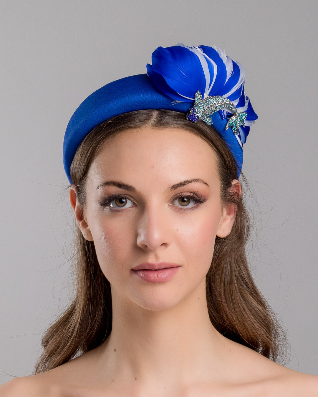 Model is showcasing the front side close up of cobalt blue high headband showcases a metal koi fish decorated with blue shades of crystals adorned with feathers behind it to create the illusion of a wave. The luxury headband is a contemporary statement. Made by Merve Bayindir Millinery.