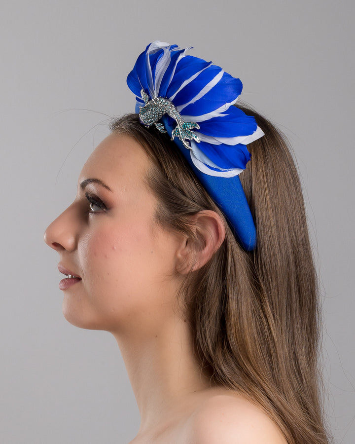Model is showcasing the left side close up of cobalt blue high headband showcases a metal koi fish decorated with blue shades of crystals adorned with feathers behind it to create the illusion of a wave. The luxury headband is a contemporary statement. Made by Merve Bayindir Millinery.