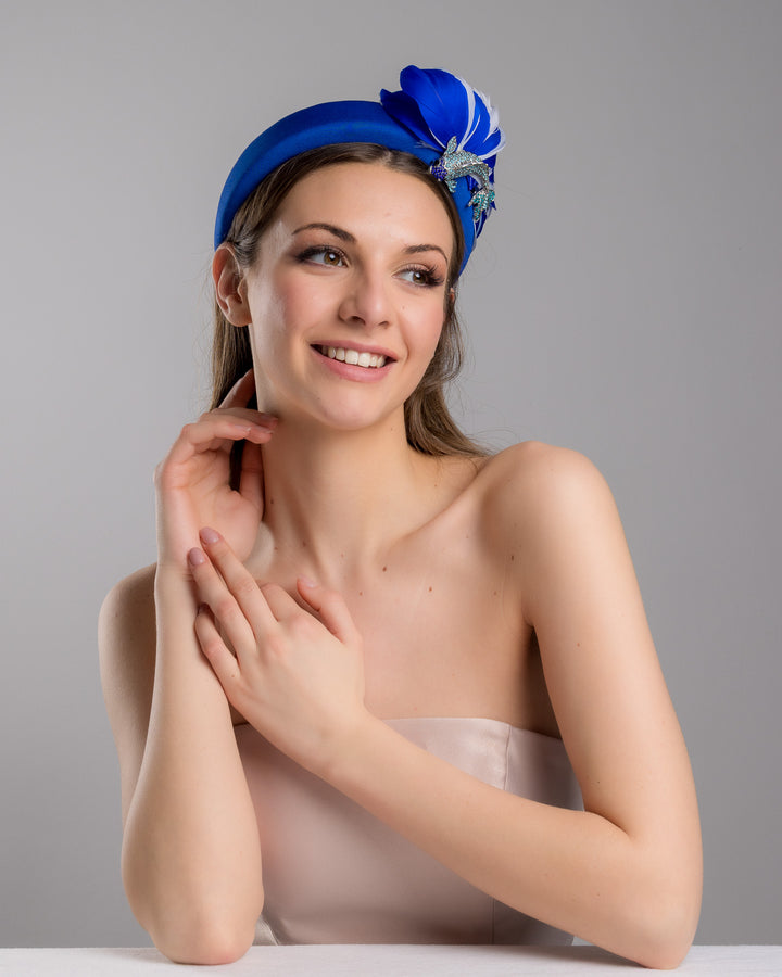 Female model is posing wearing Blue Koi cobalt blue high headband showcases a metal koi fish decorated with blue shades of crystals adorned with feathers behind it to create the illusion of a wave. The luxury headband is a contemporary statement. Made by Merve Bayindir Millinery.