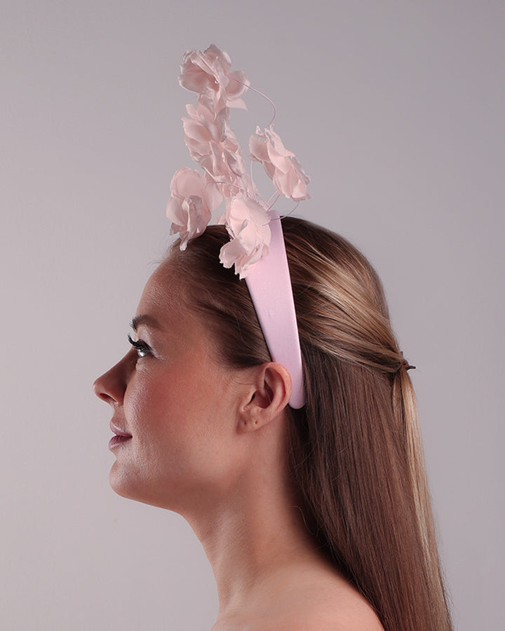 Model is showcasing the left side of a flat pink silk sateen headband decorated with pink wire branches embellished with medium pink roses centred with plastic pink pearls.
