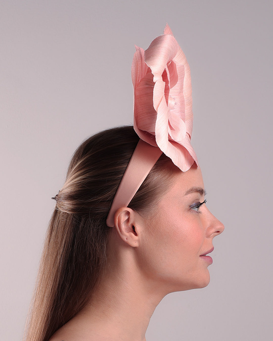 Model is showcasing the right side of a medium light pink silk abaca flower fitted on the right centre of a flat pink silk headband towards the right side of flat silk headband.