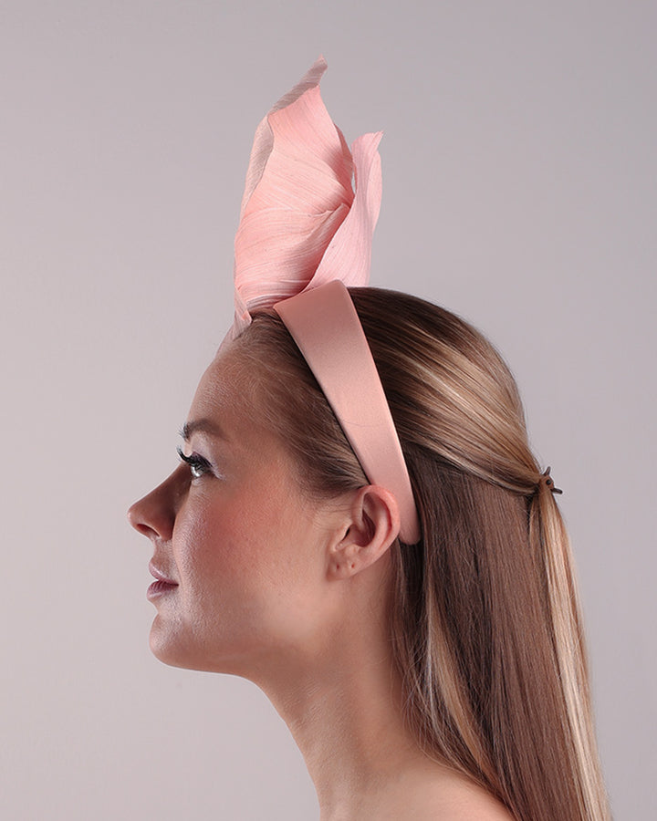 Model is showcasing the left side of a medium light pink silk abaca flower fitted on the right centre of a flat pink silk headband towards the right side of flat silk headband.