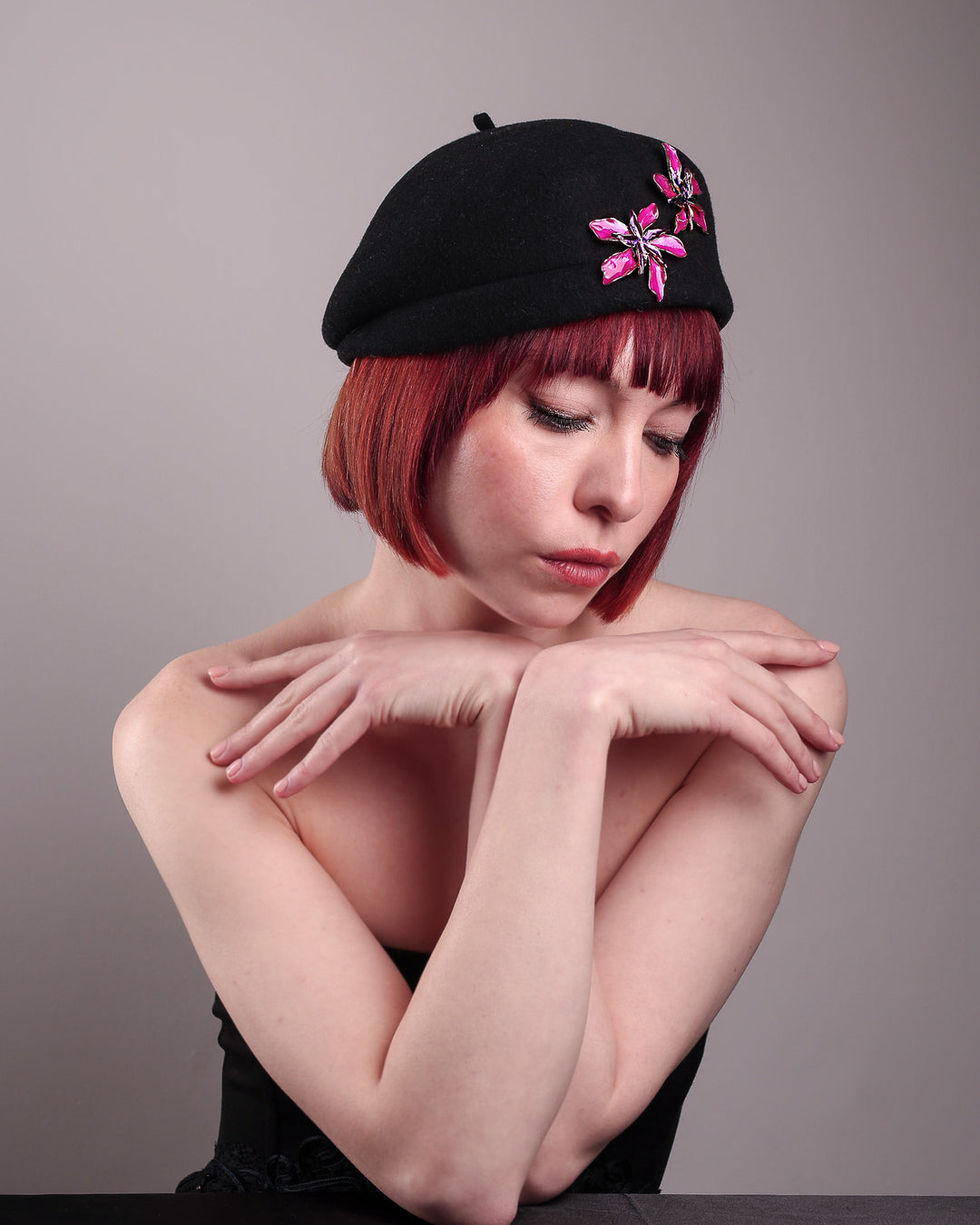 The female model is showcasing the right front side of the wool beret-style hat she is wearing. The black hat is decorated with metal, two bold floral embellishments in vivid fuchsia. The side beret is higher on the left side and shorter on the right side, with a bit of felt ribbon detail on the top centre. Designed by Merve Bayindir.