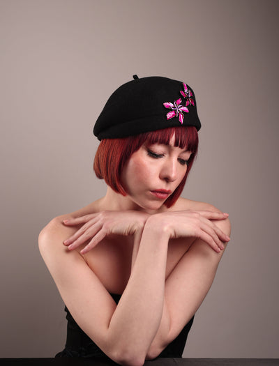 The female model is showcasing the right front side of the wool beret-style hat she is wearing. The black hat is decorated with metal, two bold floral embellishments in vivid fuchsia. The side beret is higher on the left side and shorter on the right side, with a bit of felt ribbon detail on the top centre. Designed by Merve Bayindir.
