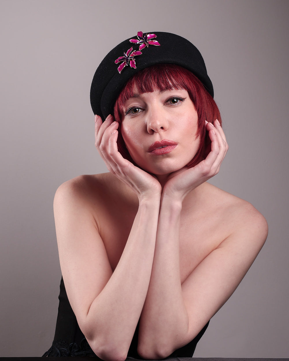The female model is showcasing the front of the wool beret-style hat she is wearing. The black hat is decorated with metal, two bold floral embellishments in vivid fuchsia. The side beret is higher on the left side and shorter on the right side, with a bit of felt ribbon detail on the top centre. Designed by Merve Bayindir.