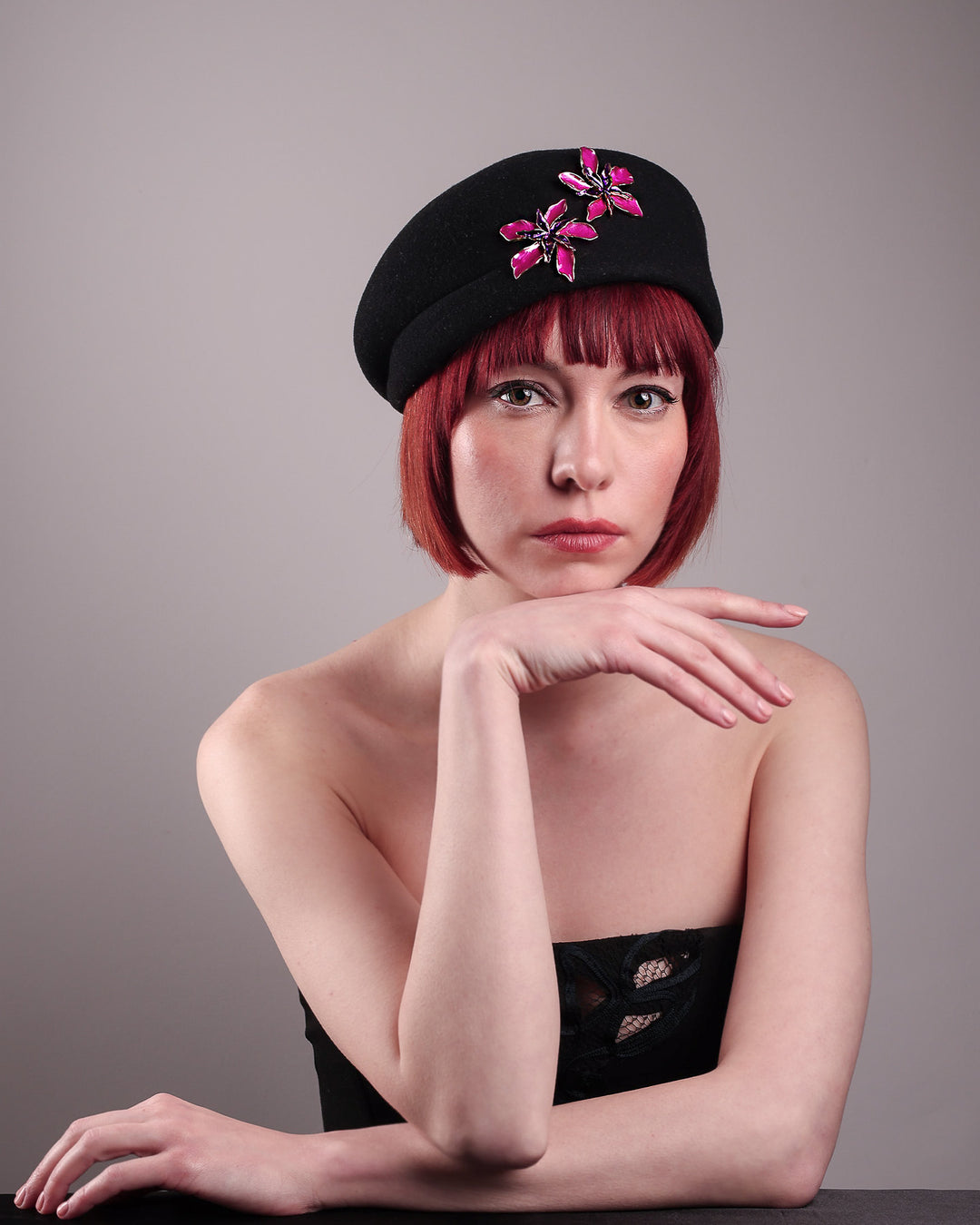 The female model is showcasing the straight front side of the wool beret-style hat she is wearing. The black hat is decorated with metal, two bold floral embellishments in vivid fuchsia. The side beret is higher on the left side and shorter on the right side, with a bit of felt ribbon detail on the top centre. Designed by Merve Bayindir.