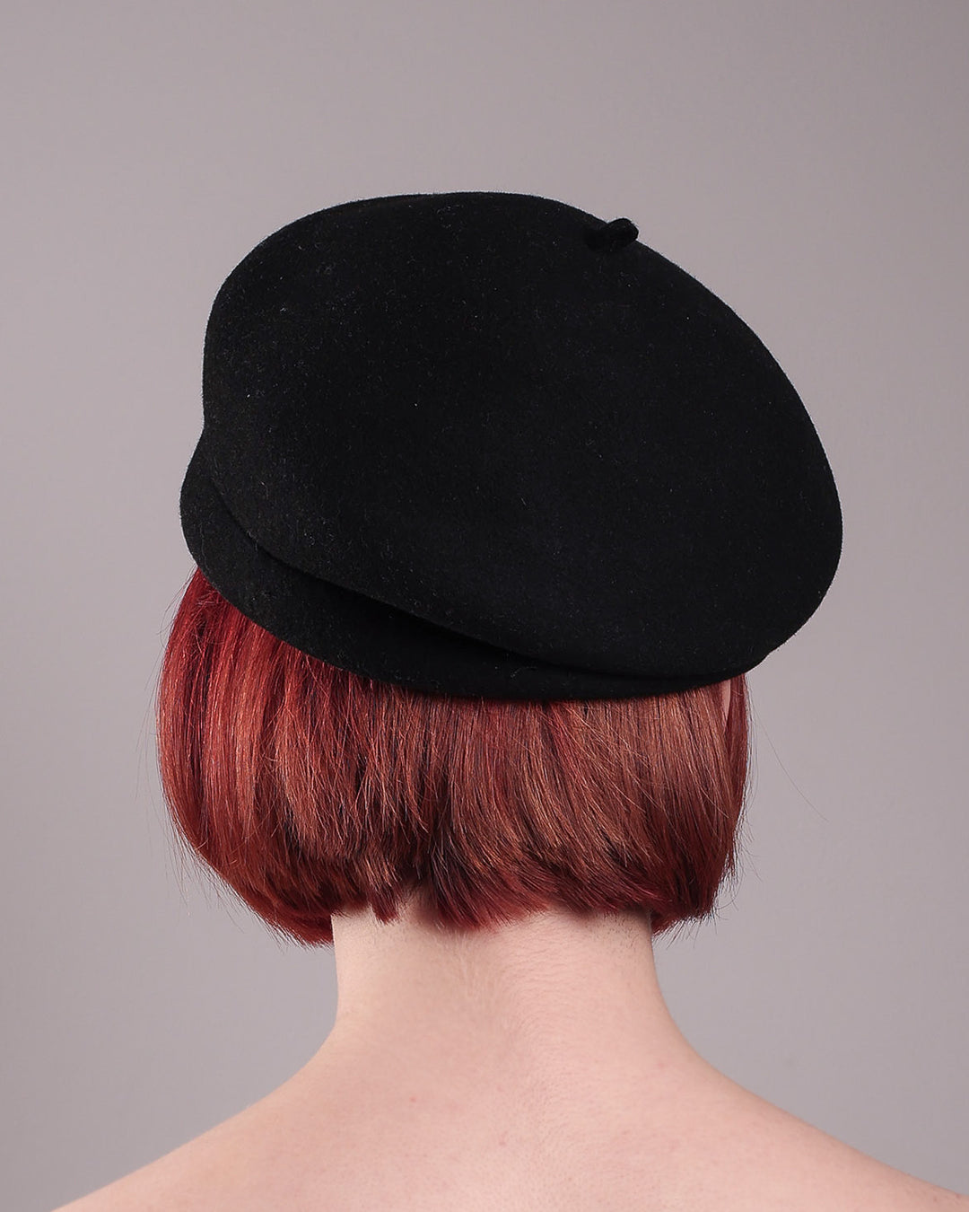 The female model is showcasing the back of the wool beret-style hat she is wearing. The black hat is decorated with metal, two bold floral embellishments in vivid fuchsia. The side beret is higher on the left side and shorter on the right side, with a bit of felt ribbon detail on the top centre. Designed by Merve Bayindir.