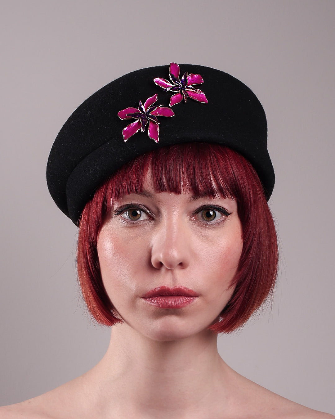 The female model is showcasing the close up of the front side of the wool beret-style hat she is wearing. The black hat is decorated with metal, two bold floral embellishments in vivid fuchsia. The side beret is higher on the left side and shorter on the right side, with a bit of felt ribbon detail on the top centre. Designed by Merve Bayindir.