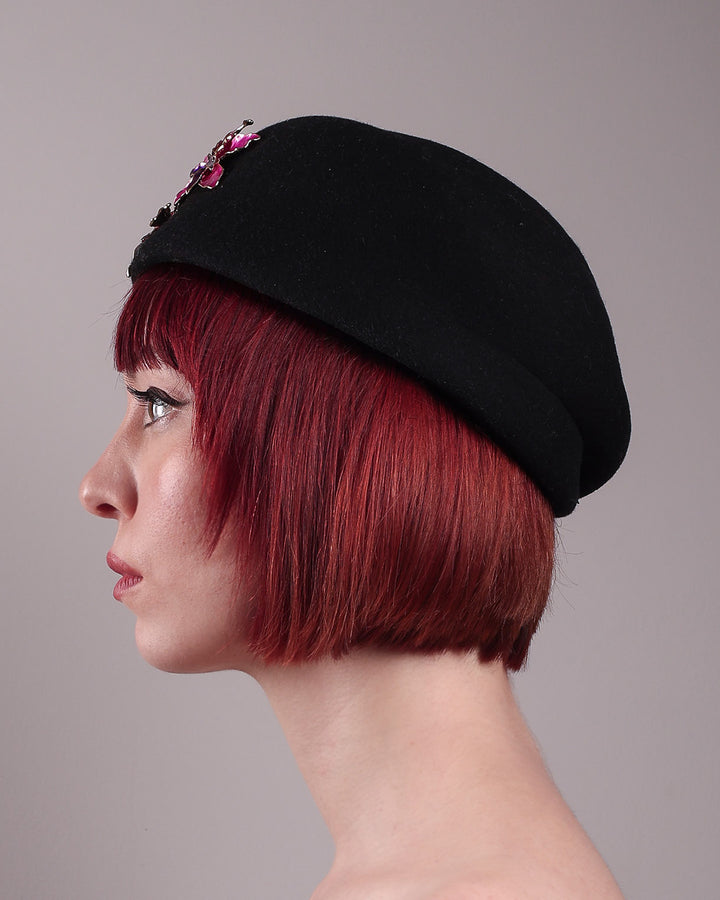 The female model is showcasing the left side close up of the wool beret-style hat she is wearing. The black hat is decorated with metal, two bold floral embellishments in vivid fuchsia. The side beret is higher on the left side and shorter on the right side, with a bit of felt ribbon detail on the top centre. Designed by Merve Bayindir.