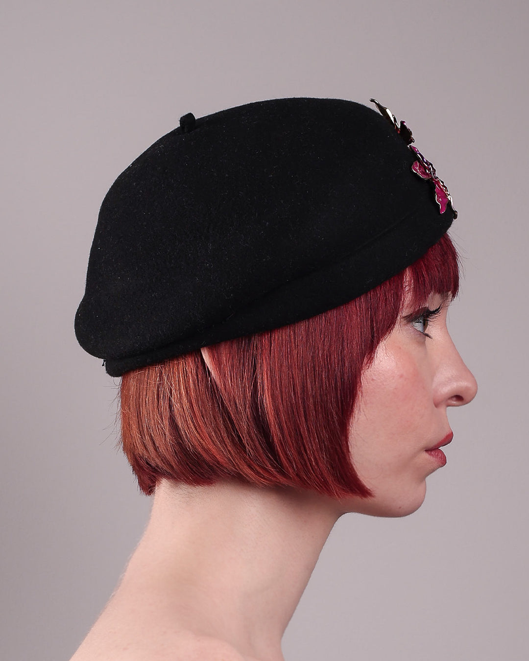 The female model is showcasing the right side close up of the wool beret-style hat she is wearing. The black hat is decorated with metal, two bold floral embellishments in vivid fuchsia. The side beret is higher on the left side and shorter on the right side, with a bit of felt ribbon detail on the top centre. Designed by Merve Bayindir.