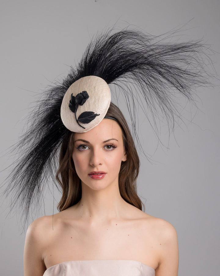 The model is showcasing the front side close up of the pink button-shaped fascinator adorned with strikingly long, burnt-black peacock feathers radiating outward, embellished with a handmade black rose. Designed and made by Merve Bayindir Millinery.
