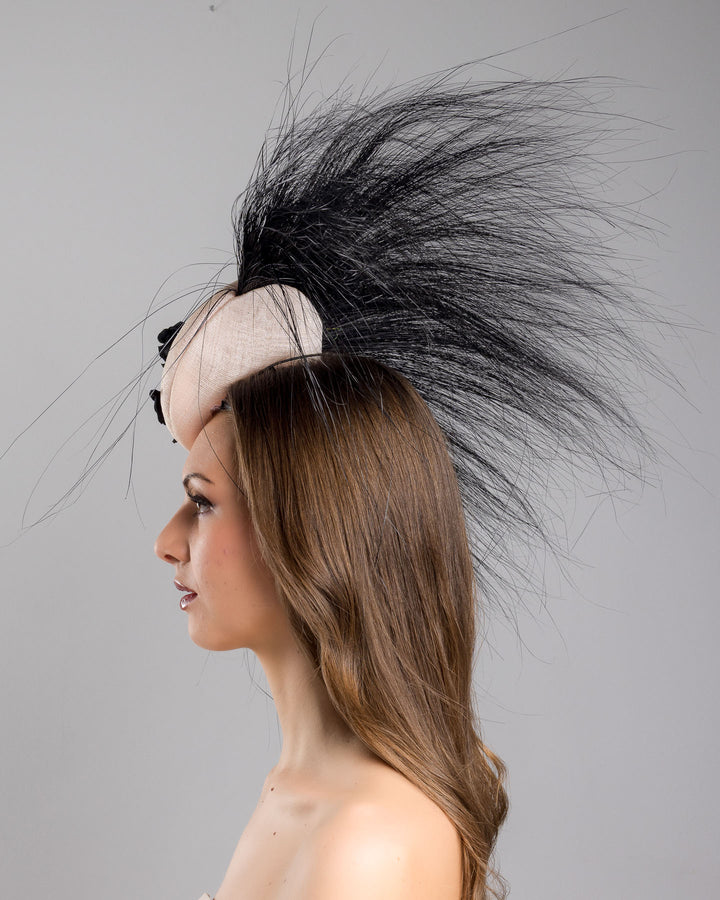 The model is showcasing the left side close up of the pink button-shaped fascinator adorned with strikingly long, burnt-black peacock feathers radiating outward, embellished with a handmade black rose. Designed and made by Merve Bayindir Millinery.
