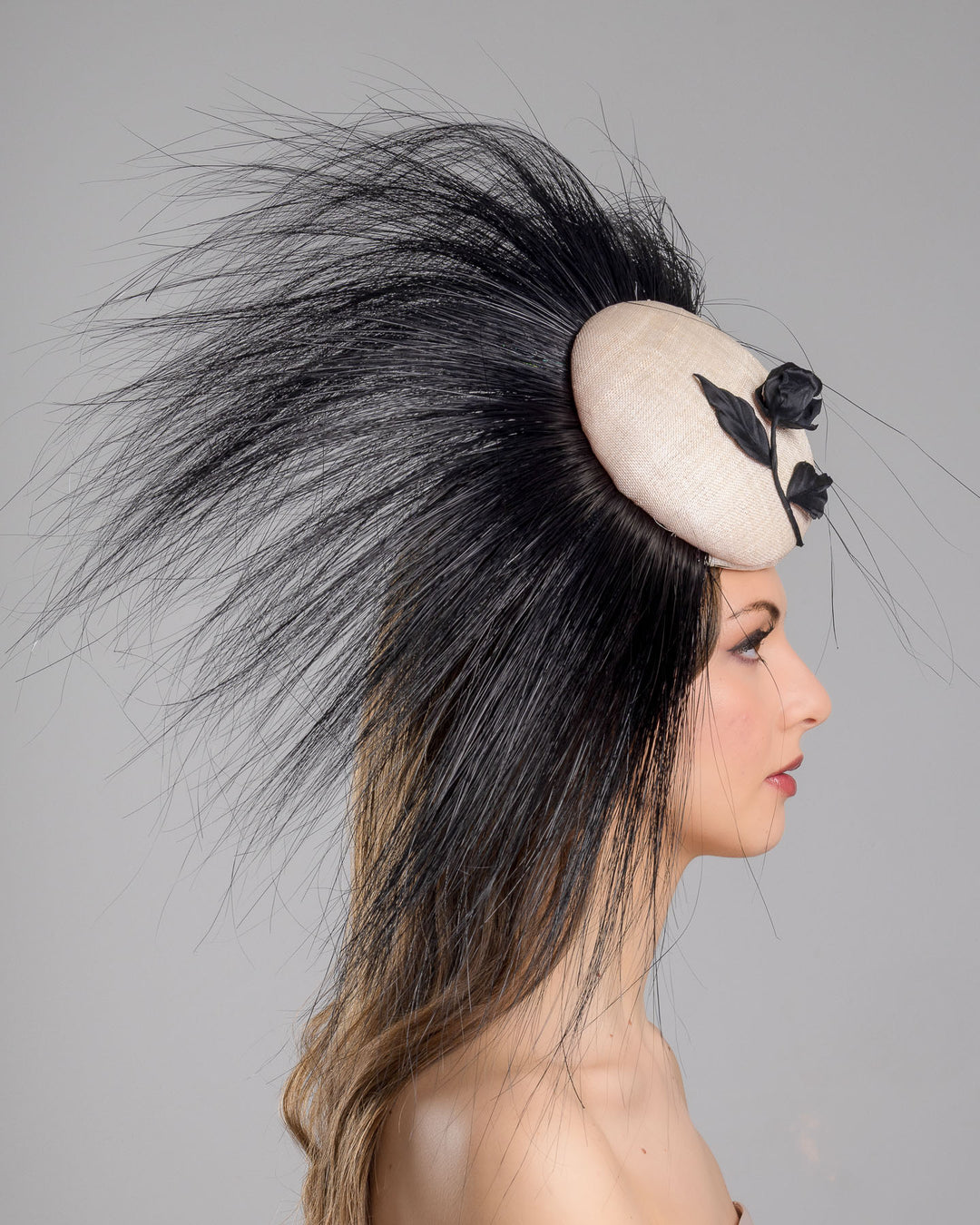 The model is showcasing the right side close up of the pink button-shaped fascinator adorned with strikingly long, burnt-black peacock feathers radiating outward, embellished with a handmade black rose. Designed and made by Merve Bayindir Millinery.
