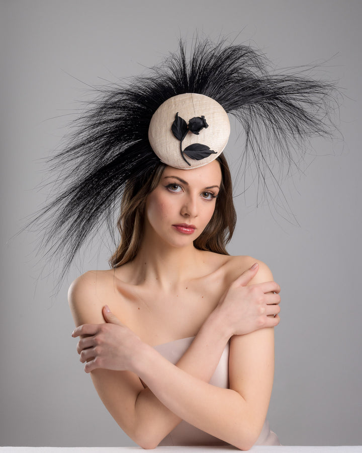 The female model is posing, wearing Briar, the pink button-shaped fascinator adorned with strikingly long, burnt-black peacock feathers radiating outward, embellished with a handmade black rose. Designed and made by Merve Bayindir Millinery.
