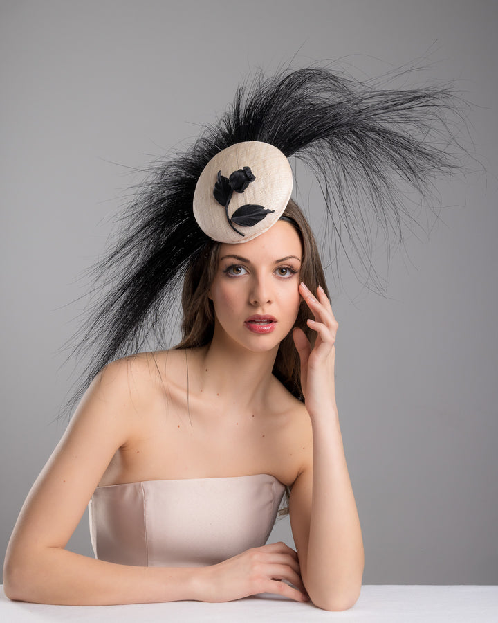 The female model is posing, wearing Briar, the pink button-shaped fascinator adorned with strikingly long, burnt-black peacock feathers radiating outward, embellished with a handmade black rose. Designed and made by Merve Bayindir Millinery.
