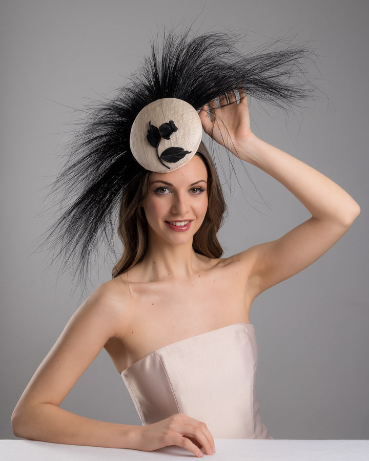The female model is posing, wearing Briar, the pink button-shaped fascinator adorned with strikingly long, burnt-black peacock feathers radiating outward, embellished with a handmade black rose. Designed and made by Merve Bayindir Millinery.
