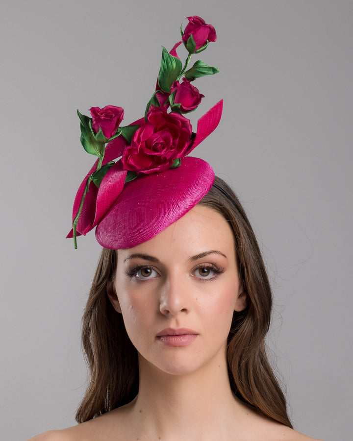 The model is showcasing the front side close up of the hot pink button-style fascinator is decorated with two long, hot pink buntal swirls surrounded by pink roses and rosebuds with green leaves. Designed and made by Merve Bayindir Millinery.
