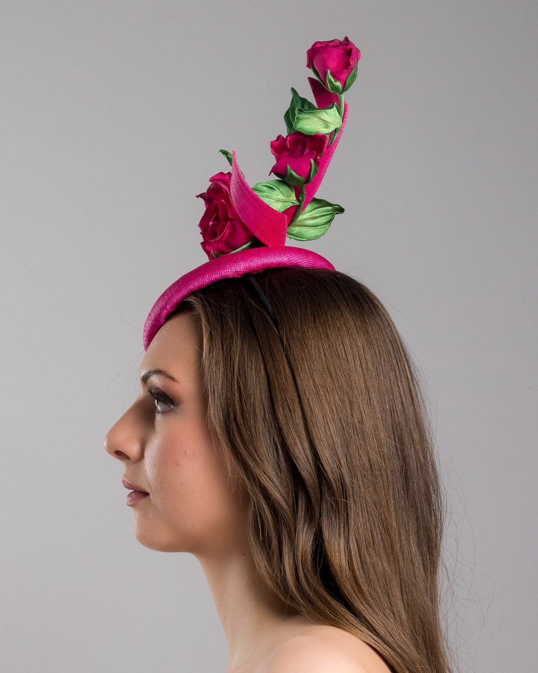 The model is showcasing the left side close up of the hot pink button-style fascinator is decorated with two long, hot pink buntal swirls surrounded by pink roses and rosebuds with green leaves. Designed and made by Merve Bayindir Millinery.
