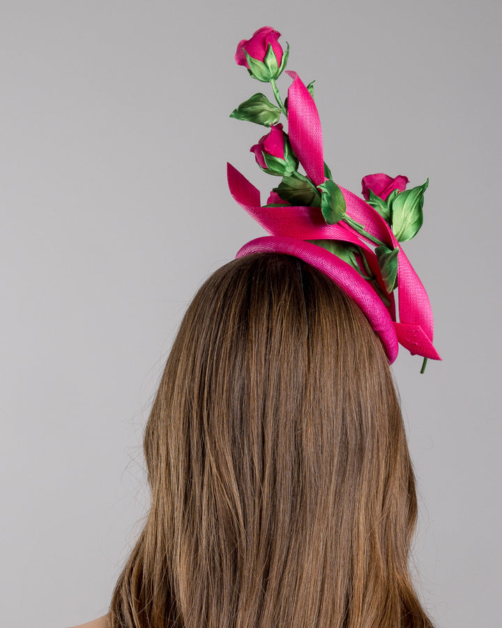 The model is showcasing the back side close up of the hot pink button-style fascinator is decorated with two long, hot pink buntal swirls surrounded by pink roses and rosebuds with green leaves. Designed and made by Merve Bayindir Millinery.

