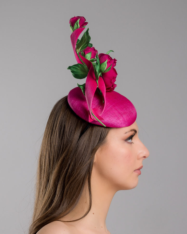 The model is showcasing the right side close up of the hot pink button-style fascinator is decorated with two long, hot pink buntal swirls surrounded by pink roses and rosebuds with green leaves. Designed and made by Merve Bayindir Millinery.
