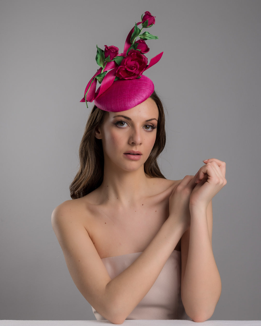 The female model is posing, wearing Brighton, the hot pink button-style fascinator is decorated with two long, hot pink buntal swirls surrounded by pink roses and rosebuds with green leaves. Designed and made by Merve Bayindir Millinery.
