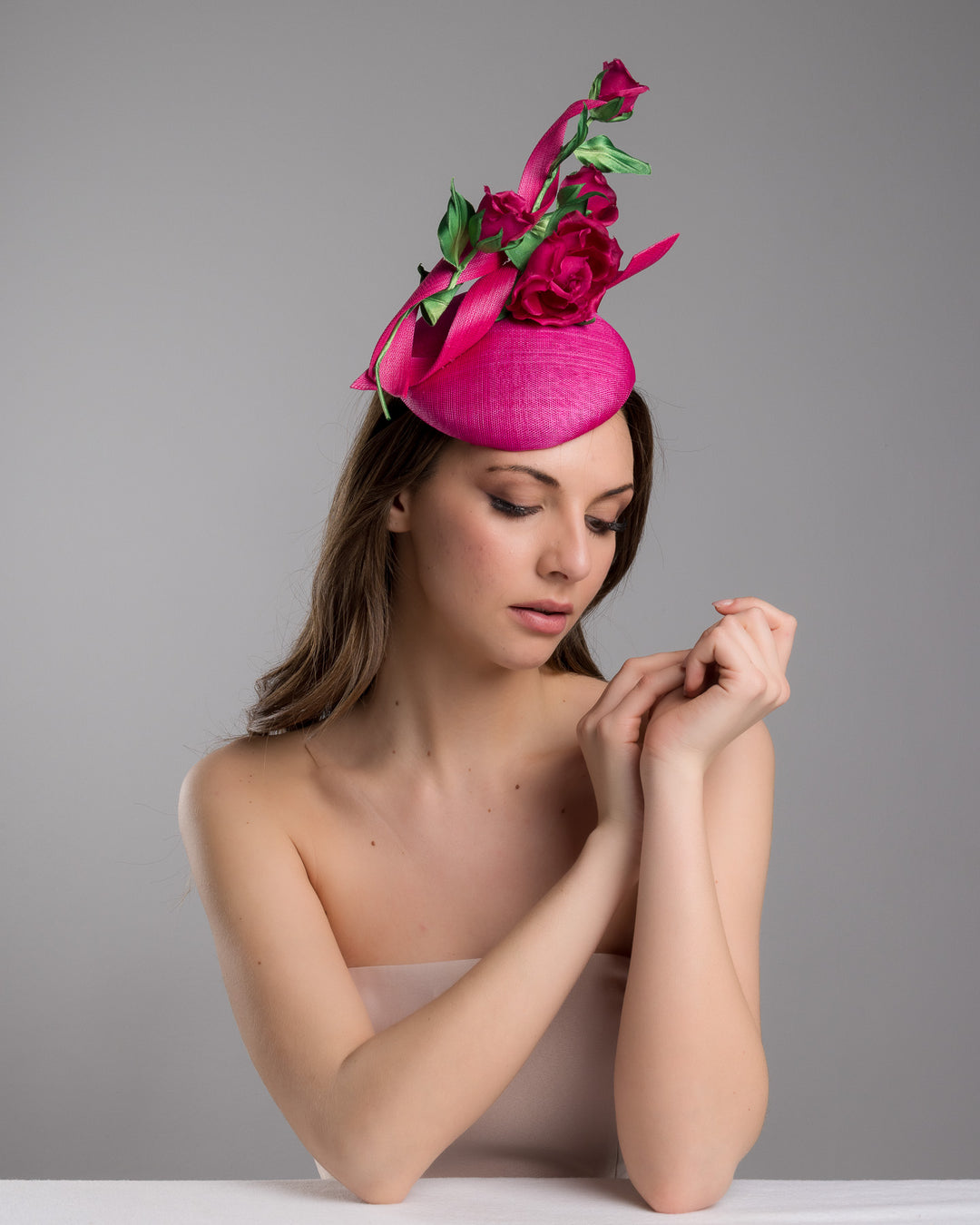 The female model is posing, wearing Brighton, the hot pink button-style fascinator is decorated with two long, hot pink buntal swirls surrounded by pink roses and rosebuds with green leaves. Designed and made by Merve Bayindir Millinery.
