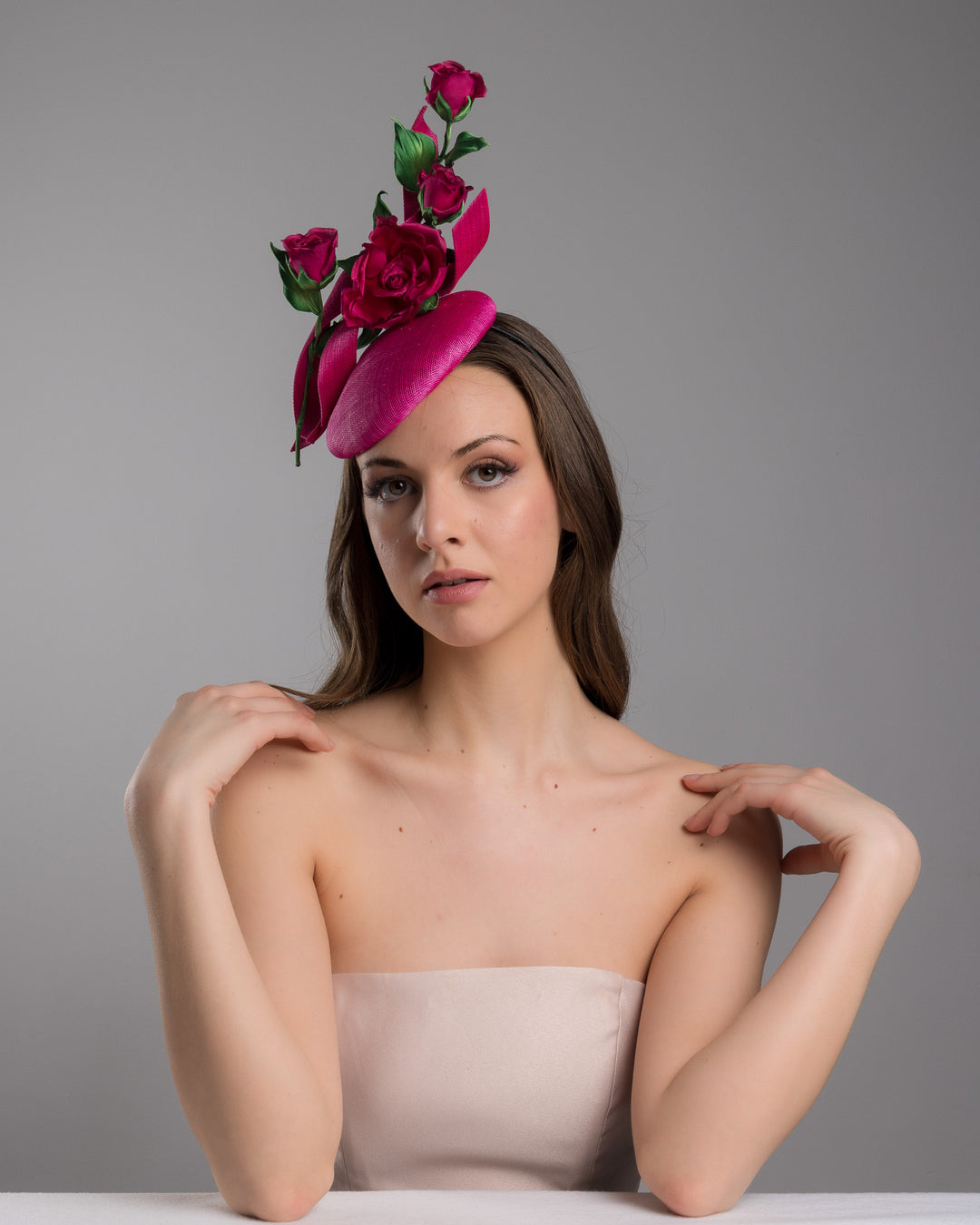 The female model is posing, wearing Brighton, the hot pink button-style fascinator is decorated with two long, hot pink buntal swirls surrounded by pink roses and rosebuds with green leaves. Designed and made by Merve Bayindir Millinery.
