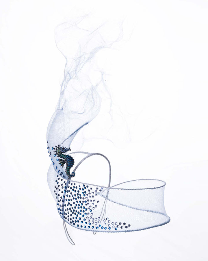 Still life picture is featuring a delicate blue mesh mask fascinator adorned with an elegant spray of light blue tulle, creating a soft, ethereal effect. The mask features intricate embellishments with blue and green crystals.