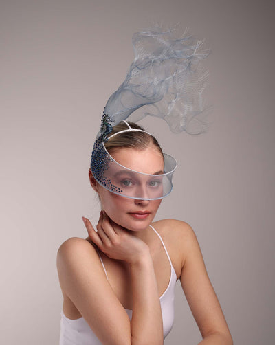 A delicate blue mesh mask fascinator adorned with an elegant spray of light blue tulle, creating a soft, ethereal effect. The mask features intricate embellishments with blue and green crystals, adding a touch of sparkle and sophistication. The model, with her hair neatly pulled back, gently touches her face, accentuating the fascinator's graceful curves and intricate details. The overall look is refined and whimsical, perfect for adding a touch of elegance to any high-fashion event.