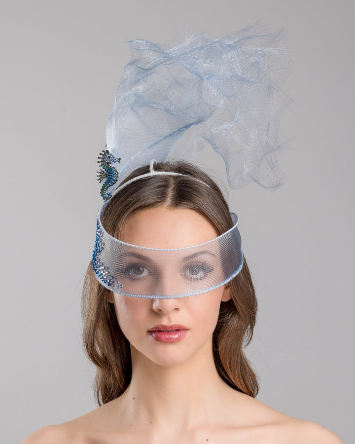 Model is showcasing the front of the delicate blue mesh mask fascinator adorned with an elegant spray of light blue tulle, creating a soft, ethereal effect. The mask features intricate embellishments with blue and green crystals.
