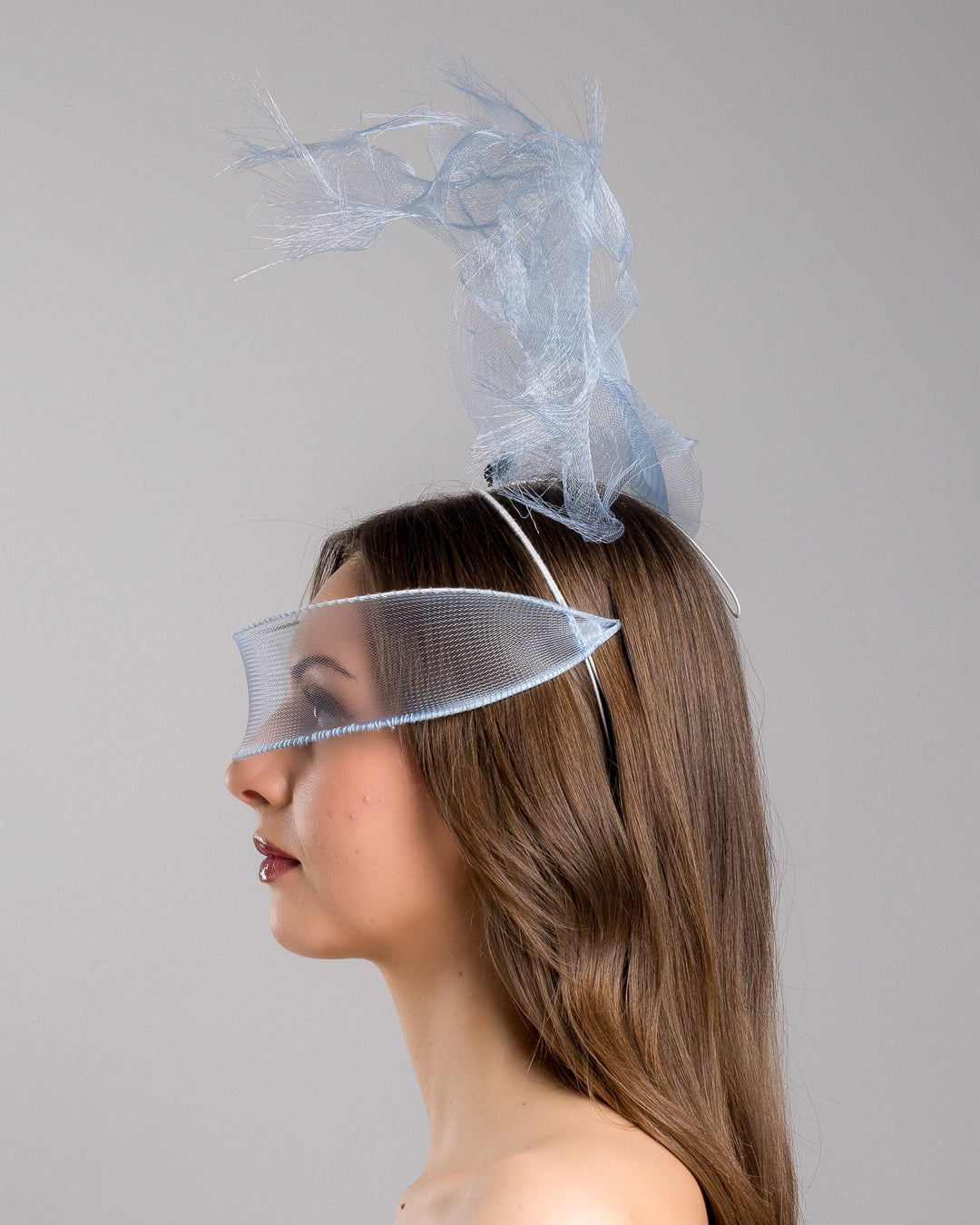 Model is showcasing the left side of the delicate blue mesh mask fascinator adorned with an elegant spray of light blue tulle, creating a soft, ethereal effect. The mask features intricate embellishments with blue and green crystals.