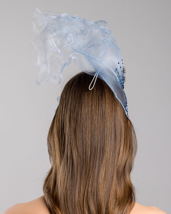 Model is showcasing the back of the delicate blue mesh mask fascinator adorned with an elegant spray of light blue tulle, creating a soft, ethereal effect. The mask features intricate embellishments with blue and green crystals.