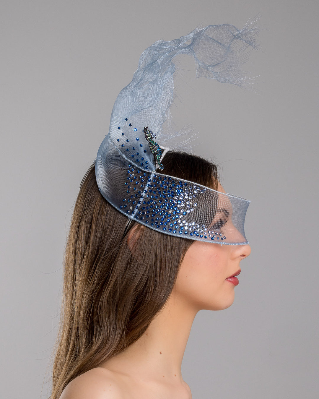 Model is showcasing the right side of the delicate blue mesh mask fascinator adorned with an elegant spray of light blue tulle, creating a soft, ethereal effect. The mask features intricate embellishments with blue and green crystals.
