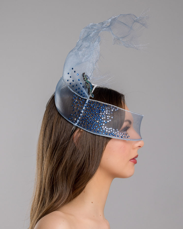Model is showcasing the right side of the delicate blue mesh mask fascinator adorned with an elegant spray of light blue tulle, creating a soft, ethereal effect. The mask features intricate embellishments with blue and green crystals.