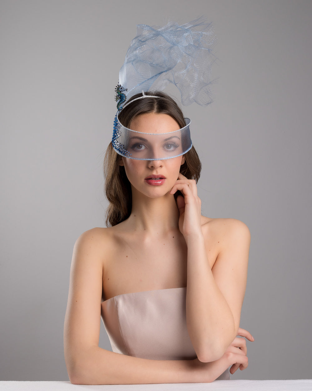 Model is posing wearing couture Merve Bayindir Brizo mask, showcasing the delicate blue mesh mask fascinator adorned with an elegant spray of light blue tulle, creating a soft, ethereal effect. The mask features intricate embellishments with blue and green crystals.
