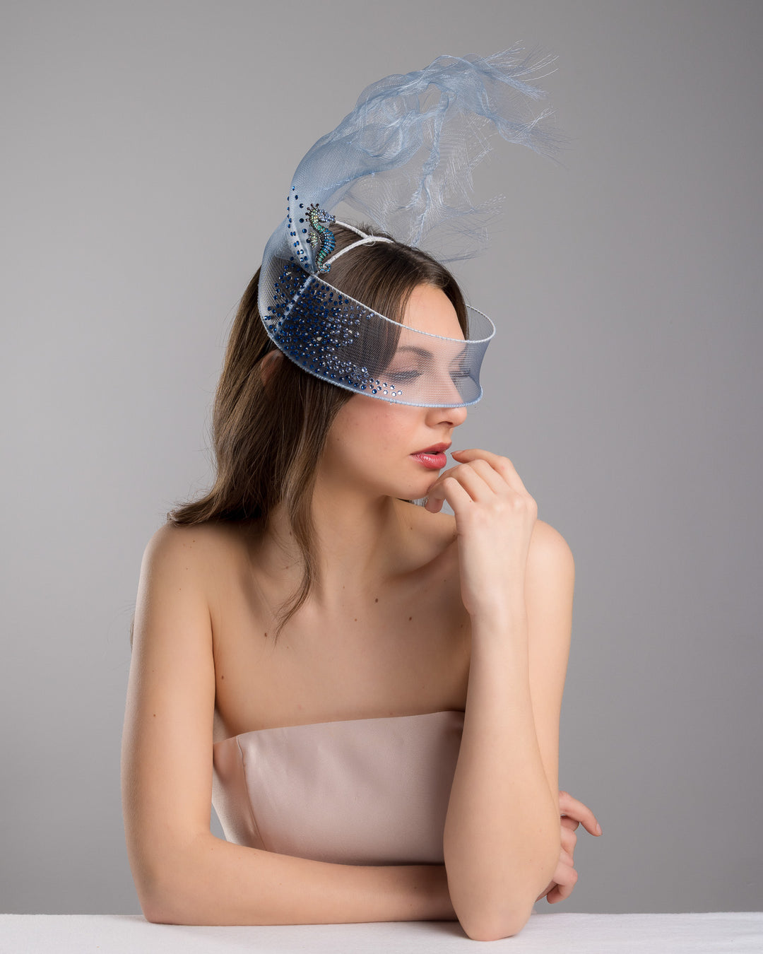 Model is posing wearing couture Merve Bayindir Brizo mask, showcasing the delicate blue mesh mask fascinator adorned with an elegant spray of light blue tulle, creating a soft, ethereal effect. The mask features intricate embellishments with blue and green crystals.