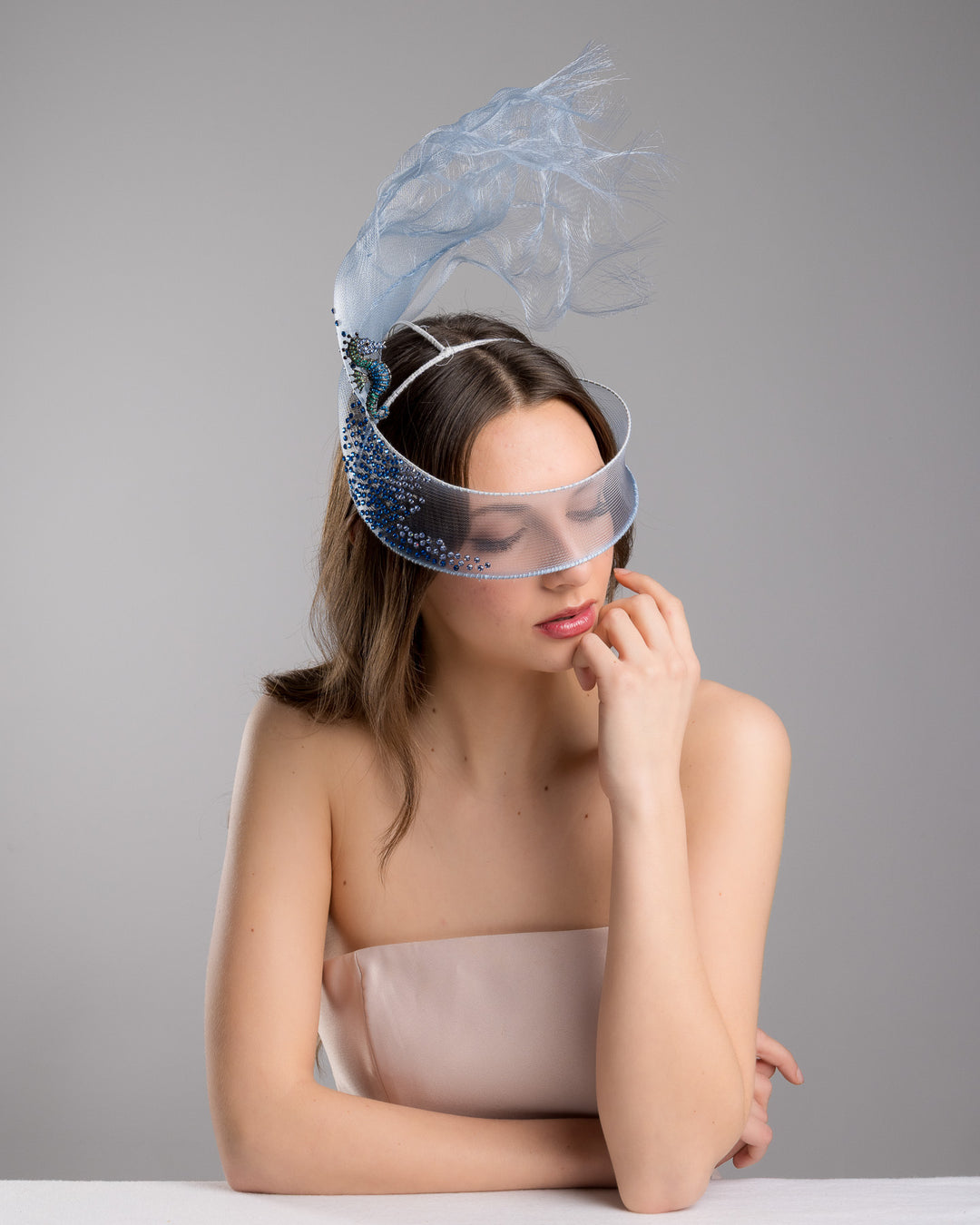 Model is posing wearing couture Merve Bayindir Brizo mask, showcasing the delicate blue mesh mask fascinator adorned with an elegant spray of light blue tulle, creating a soft, ethereal effect. The mask features intricate embellishments with blue and green crystals.
