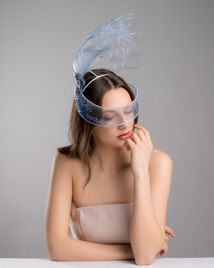 Model is posing wearing couture Merve Bayindir Brizo mask, showcasing the delicate blue mesh mask fascinator adorned with an elegant spray of light blue tulle, creating a soft, ethereal effect. The mask features intricate embellishments with blue and green crystals.