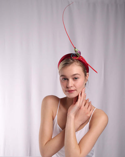 The model is wearing a striking bright red headband adorned with a multi-colored crystal-embellished metal brooch in the shape of a chameleon, adding a whimsical and playful touch. The headband is further accentuated by a long, elegant red quill that extends upwards, creating a dramatic and eye-catching effect. 