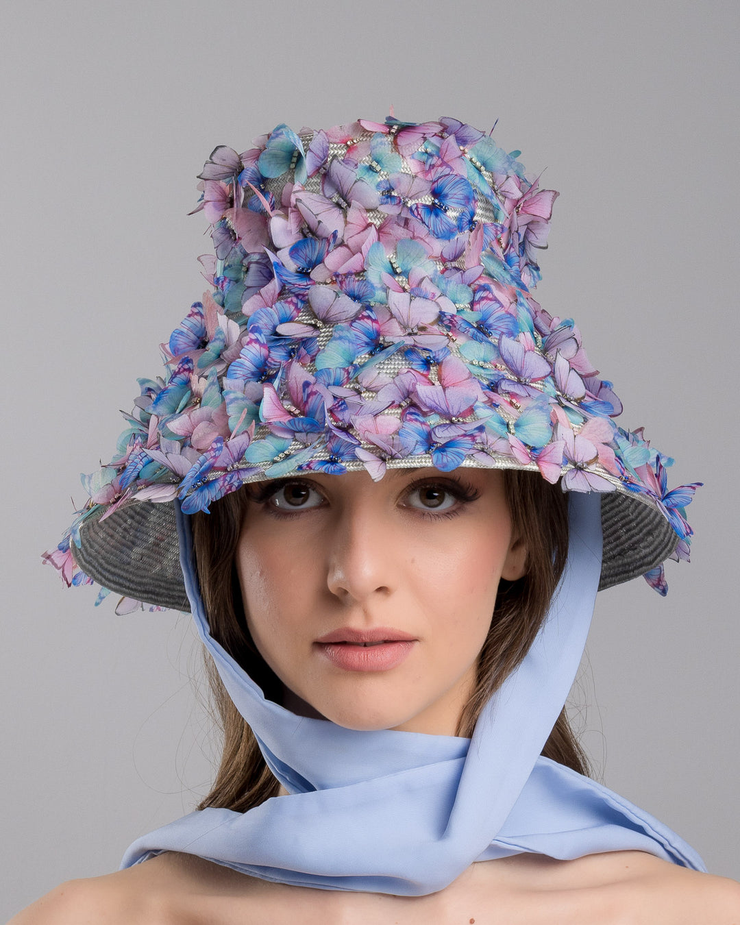 The model showcases the front side of the   high fashion blue bucket hat  with long blue silk chiffon ribbons dropping on the sides, the hat is adorned with multi colour organza butterflies. Designed and made by Merve Bayindir Millinery.