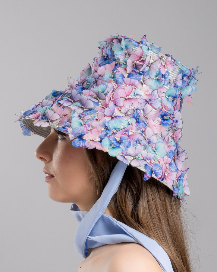 The model showcases the left side of the   high fashion blue bucket hat  with long blue silk chiffon ribbons dropping on the sides, the hat is adorned with multi colour organza butterflies. Designed and made by Merve Bayindir Millinery.